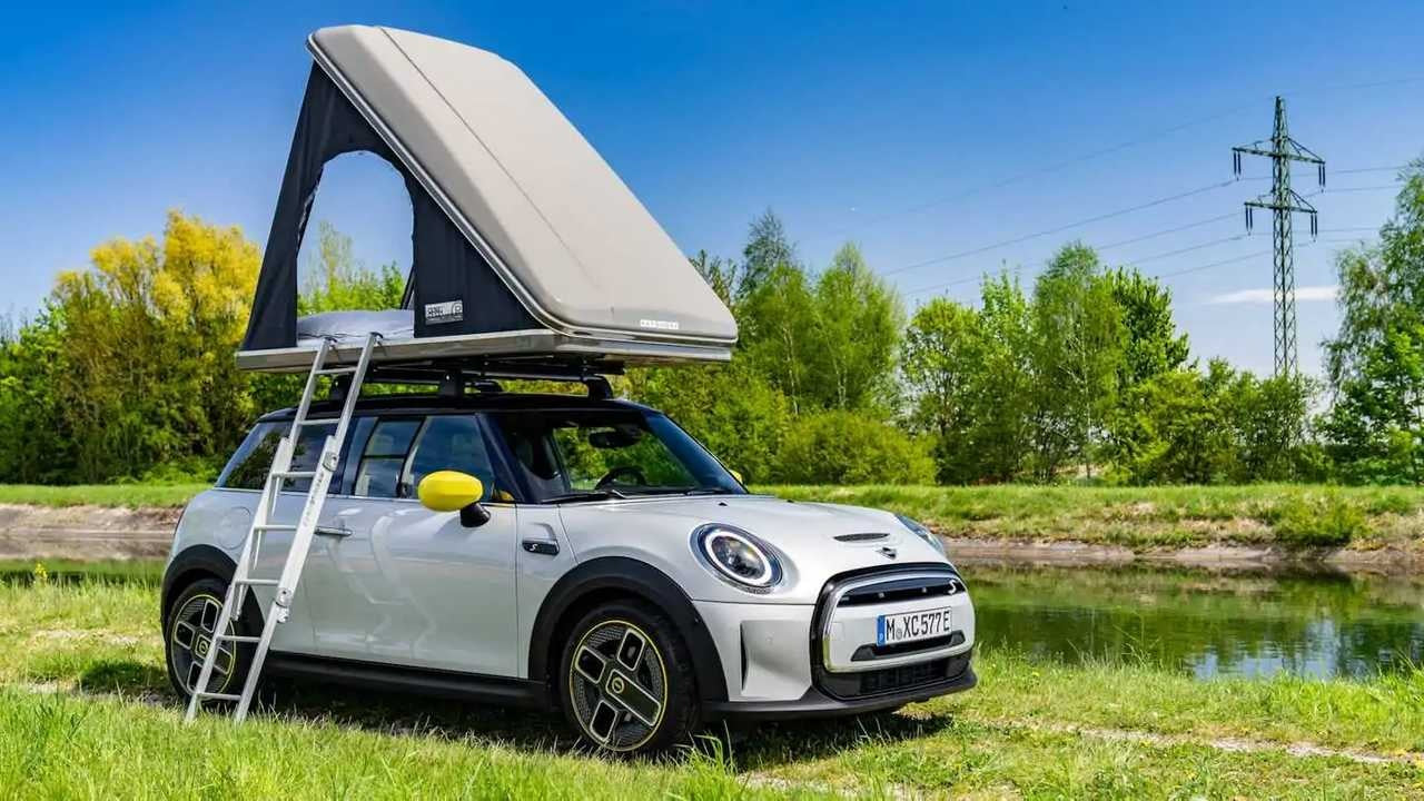 White car with a rooftop tent parked in a grassy area with trees and a clear blue sky.